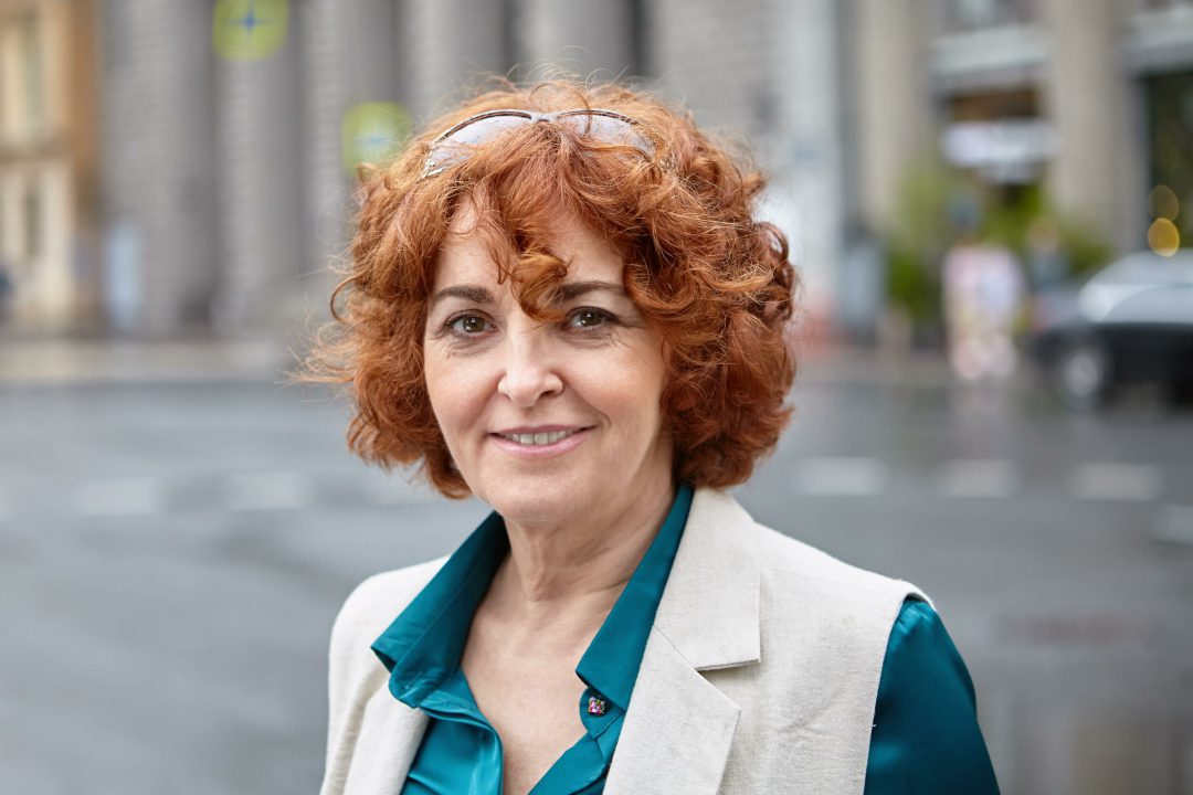 Smiling mature brown-haired woman in front of a city street in rainy weather. Senior white female close-up portrait outdoors.